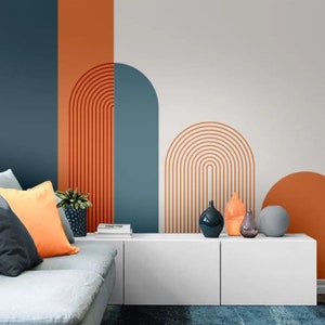 Op de afbeelding: Een woonkamer interieurontwerp met een witte muur met een geometrisch patroon van oranje en blauwe bogen. De muur is geaccentueerd met een wit zwevend rek en een grijze bank met oranje en grijze kussens.