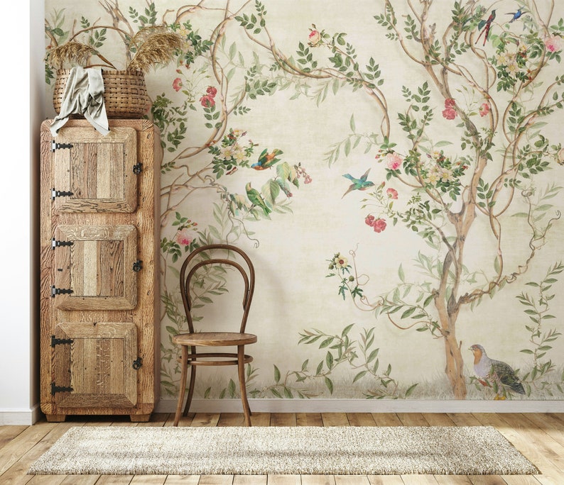 May include: A rustic wooden cabinet with three doors stands next to a wooden chair and a floral wallpaper. A woven basket with dried plants and a folded cloth sits atop the cabinet. The wallpaper features a tree with birds and flowers.