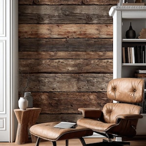 Puede incluir: Un salón acogedor con un sillón de cuero marrón y un reposapiés, frente a una pared de tablones de madera rústica. Una estantería blanca y una pequeña mesa auxiliar de madera con un jarrón completan el interior.