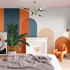 Op de afbeelding: Een slaapkamerinterieur met een wit bed, een witte open haard en een muur met een abstract geometrisch ontwerp in oranje, blauw en wit. De muur heeft een reeks bogen en lijnen. Een grote groene plant staat in een pot op de vloer. Een moderne oranje stoel staat in de hoek van de kamer.