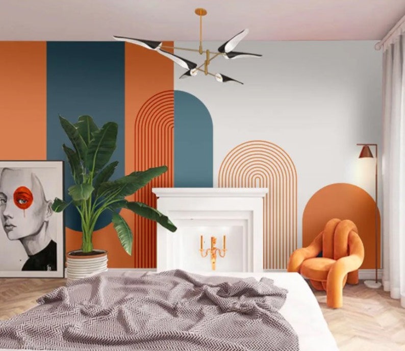 Op de afbeelding: Een slaapkamerinterieur met een wit bed, een witte open haard en een muur met een abstract geometrisch ontwerp in oranje, blauw en wit. De muur heeft een reeks bogen en lijnen. Een grote groene plant staat in een pot op de vloer. Een moderne oranje stoel staat in de hoek van de kamer.