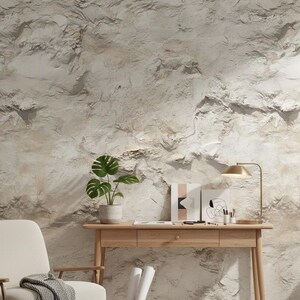 May include: A light wood desk with a gold lamp, potted plant, and artwork against a textured, light-colored wall. A woven basket holds rolled papers, and a chair with a gray blanket is nearby.