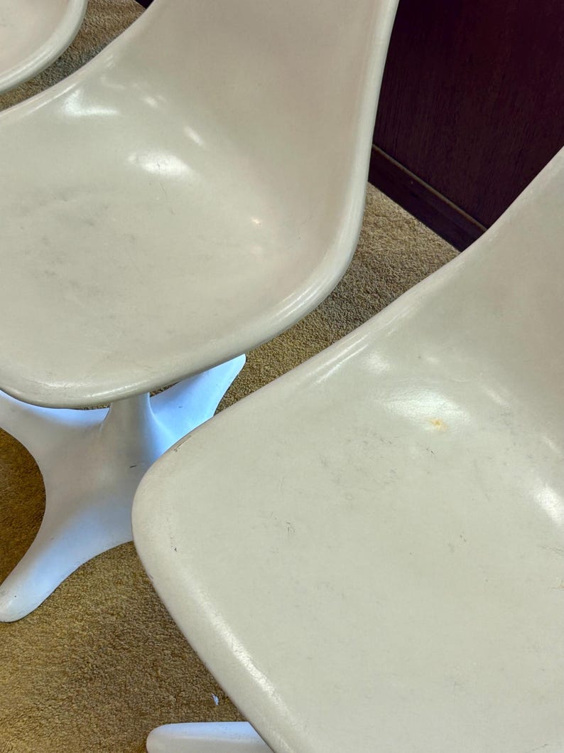 Pode incluir: Tr&ecirc;s cadeiras de pl&aacute;stico branco com base em forma de estrela. As cadeiras est&atilde;o dispostas em fila sobre um tapete bege.