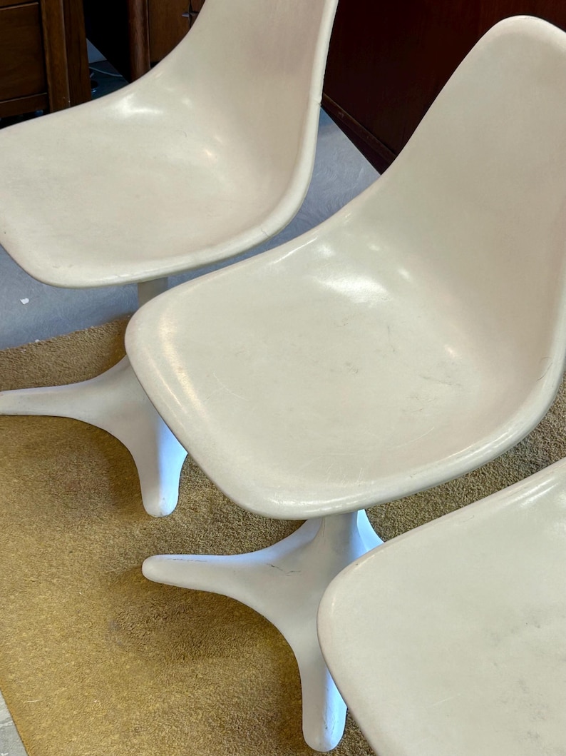 Pode incluir: Tr&ecirc;s cadeiras brancas de fibra de vidro com uma base &uacute;nica em forma de estrela. As cadeiras est&atilde;o dispostas em fila, com a base de cada cadeira vis&iacute;vel.