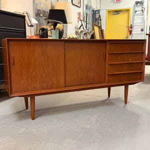 May include: A rectangular wooden sideboard featuring a warm brown wood grain finish. The piece includes two sliding doors with vertical handles and a column of four horizontal drawers on the right side, all supported by four tapered wooden legs.
