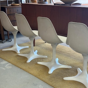 Pode incluir: Quatro cadeiras brancas em forma de tulipa com uma base em forma de estrela est&atilde;o dispostas em fila sobre um tapete bege. As cadeiras est&atilde;o em frente a uma mesa de madeira escura.