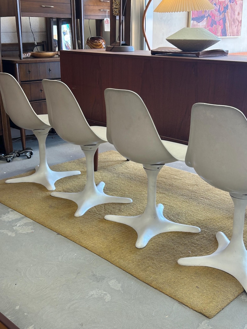Pode incluir: Quatro cadeiras brancas em forma de tulipa com uma base em forma de estrela est&atilde;o dispostas em fila sobre um tapete bege. As cadeiras est&atilde;o em frente a uma mesa de madeira escura.