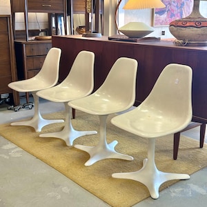 Pode incluir: Quatro cadeiras brancas em forma de tulipa com base em forma de estrela, dispostas em fila sobre um tapete bege. As cadeiras est&atilde;o em uma sala com uma mesa de madeira e outros m&oacute;veis.