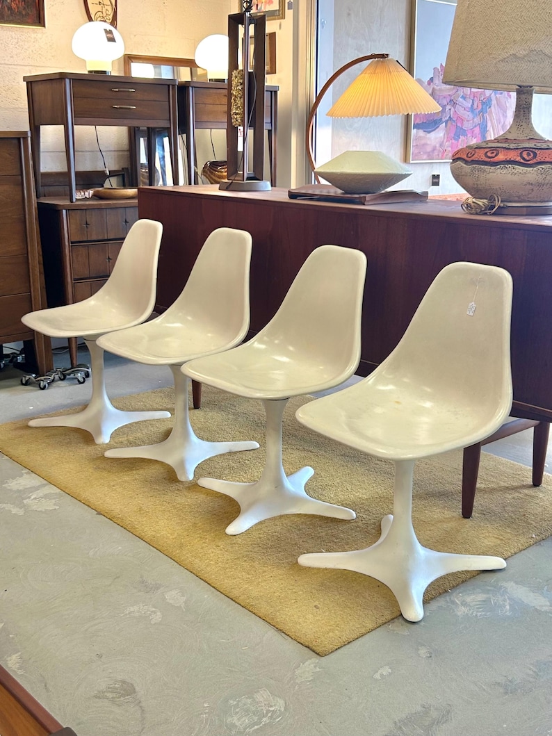 Pode incluir: Quatro cadeiras brancas em forma de tulipa com base em forma de estrela, dispostas em fila sobre um tapete bege. As cadeiras est&atilde;o em uma sala com uma mesa de madeira e outros m&oacute;veis.