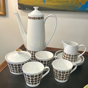 Może przedstawiać: Biały ceramiczny zestaw do herbaty z geometrycznym wzorem w kolorze brązowym i złotym. Zestaw zawiera czajnik, dzbanek do śmietanki, cukiernicę i cztery filiżanki i spodki.