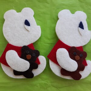 May include: Two felt ornaments shaped like bears. The bears are white with red shirts and are holding brown teddy bears.