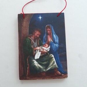 Puede incluir: Un adorno navideño de madera que representa la escena de la Natividad. El adorno muestra a María, José y el niño Jesús en un establo con una estrella sobre ellos. El adorno está pintado en un estilo realista con colores cálidos.