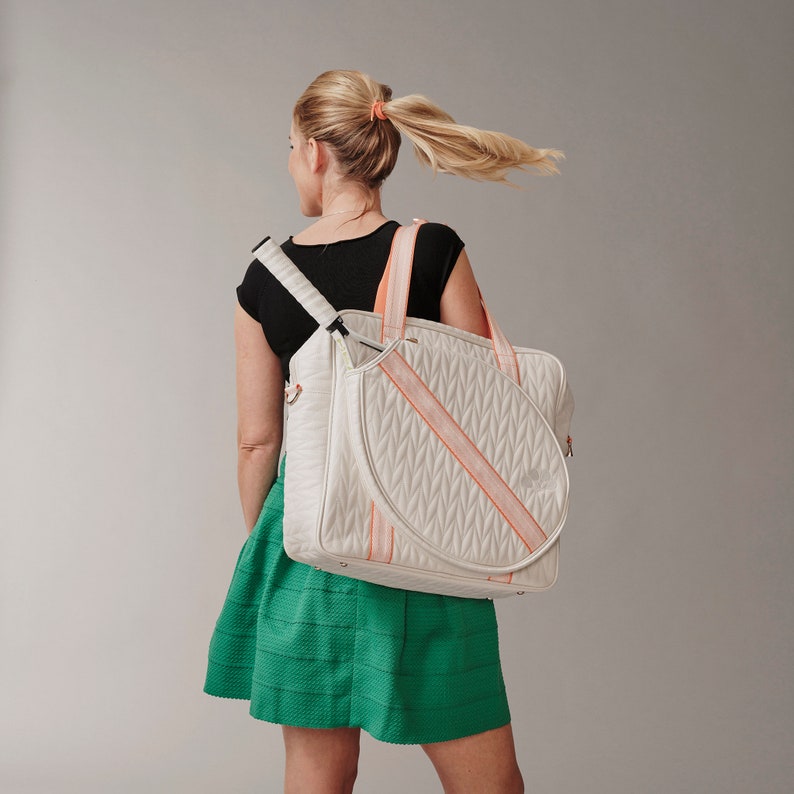 Woman holding a tennis bag in quilted white vegan leather with orange webbing and solid golden metal pullers over her shoulder.