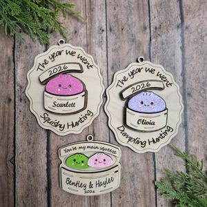 Puede incluir: Tres adornos de madera con diseños en capas de dumplings en vaporeras de bambú. Dos adornos grandes festoneados dicen 'The year we went 2026 Squishy Hunting' con 'Scarlett' y 'Dumpling Hunting' con 'Olivia'. Un adorno más pequeño dice 'You're my main squeeze' con 'Bentley & Haylee 2026'.