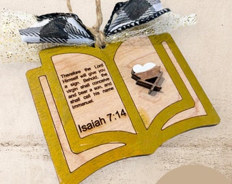 May include: Wooden ornament in the shape of an open book, with the text "Isaiah 7:14" and a Bible verse. The ornament has a gold border and a small wooden nativity scene. It is adorned with a black and white plaid bow and gold glitter ribbon.