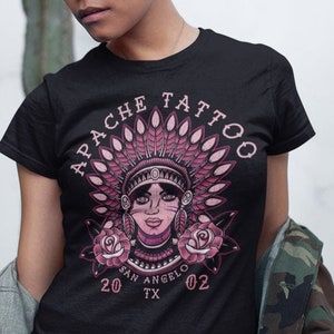Può includere: Una maglietta nera con una grafica rosa di una donna nativa americana che indossa un copricapo e circondata da rose. Il testo "APACHE TATTOO" è sopra la grafica e "SAN ANGELO TX 20 02" è sotto la grafica.