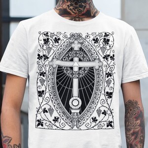 May include: White t-shirt featuring a detailed black and white graphic of a cross within an oval frame, surrounded by floral and vine motifs. The design includes intricate details and text. The shirt is a classic crew neck style.