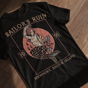 May include: Black t-shirt with a vintage-style graphic of a mermaid with a flower and the text "Sailor's Ruin 19 91 Newport, Rhode Island".