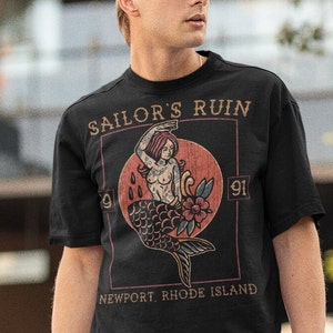 May include: A black t-shirt with a vintage-style graphic of a mermaid with the text "Sailor's Ruin" and "Newport, Rhode Island".