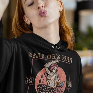 May include: Black hoodie with a vintage-style graphic of a mermaid with a floral design. The graphic is surrounded by a rectangle with the text "Sailor's Ruin" and the numbers "19" and "91". The text "Newport, Rhode Island" is at the bottom of the graphic.