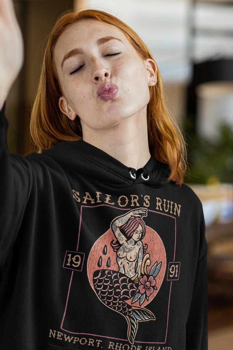 May include: Black hoodie with a vintage-style graphic of a mermaid with a floral design. The graphic is surrounded by a rectangle with the text "Sailor's Ruin" and the numbers "19" and "91". The text "Newport, Rhode Island" is at the bottom of the graphic.