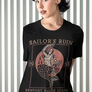 May include: Black t-shirt with a vintage-style graphic of a mermaid with tattoos. The graphic is in a square frame with the text "Sailor's Ruin 19 91 Newport, Rhode Island".