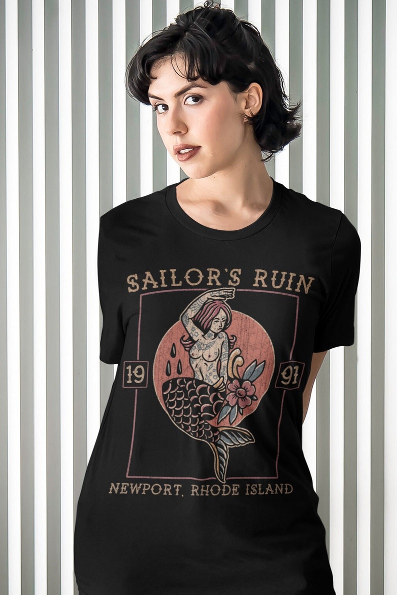 May include: Black t-shirt with a vintage-style graphic of a mermaid with tattoos. The graphic is in a square frame with the text "Sailor's Ruin 19 91 Newport, Rhode Island".