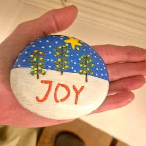 May include: A hand-painted, round stone with a winter scene. The top half features a blue sky with white dots and a yellow star. Three Christmas trees are below the star. The bottom half is white with the word "JOY" in orange.
