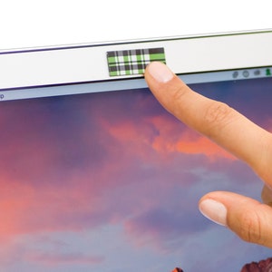 May include: A laptop computer with a black and white plaid webcam cover over the camera lens. The laptop screen displays a pink and purple cloudy sky with a mountain range in the distance.