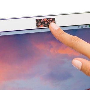 May include: A close-up of a laptop computer with a floral patterned webcam cover. The cover is black with pink and white flowers. A finger is shown pushing the cover down to reveal the webcam.
