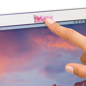 May include: A pink and white patterned webcam cover is placed over the camera lens of a laptop. A finger is shown touching the cover.