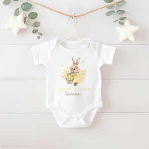 May include: White baby onesie with a watercolor illustration of a bunny holding a basket of eggs, wearing a yellow tutu, and surrounded by yellow flowers. The text "HAPPY EASTER Sienna" is printed in gold.