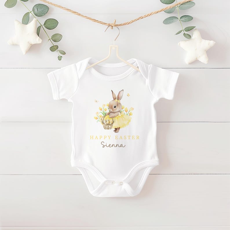 May include: White baby onesie with a watercolor illustration of a bunny holding a basket of eggs, wearing a yellow tutu, and surrounded by yellow flowers. The text "HAPPY EASTER Sienna" is printed in gold.