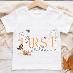 May include: A white short-sleeved t-shirt featuring the text My First Halloween in orange and black script. The graphic includes illustrations of two small ghosts, one wearing a witch hat, two pumpkins, a bat, and a spider hanging from a thread.