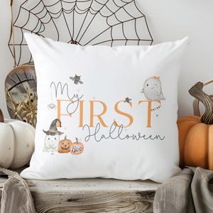 May include: A white square throw pillow featuring a printed graphic with the text My FIRST Halloween in orange and black script. The design includes illustrations of two small ghosts, a pumpkin with the word Boo, a bat, and a spider.