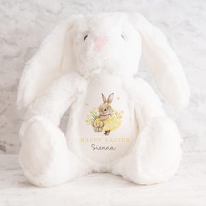 May include: A white plush bunny with long ears and a pink nose. The bunny has a printed design on its chest featuring a rabbit in a yellow dress, a basket, and the text "HAPPY EASTER" and a name. A soft, cuddly Easter-themed toy.