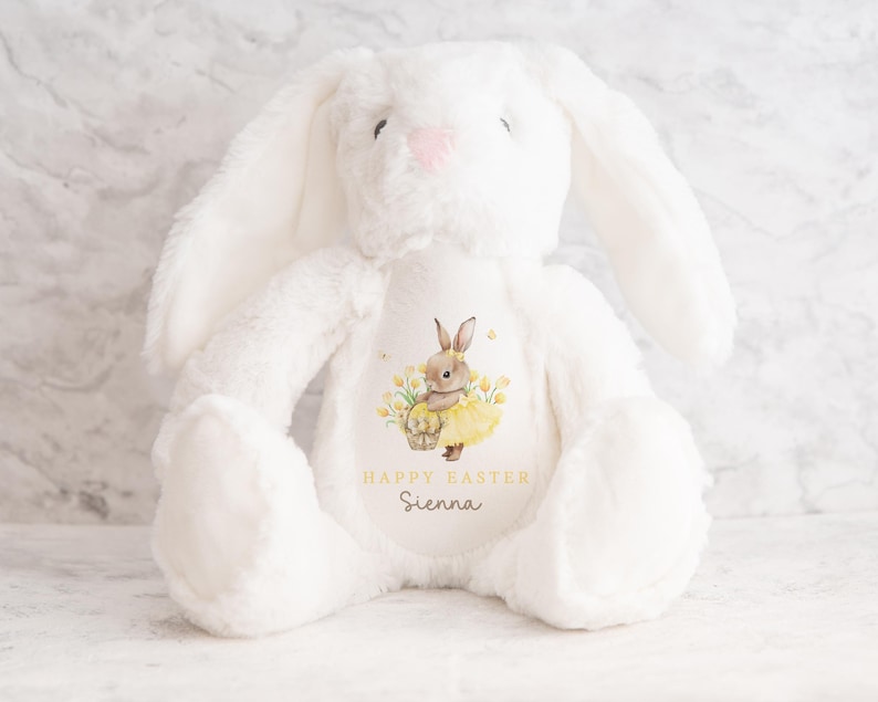 May include: A white plush bunny with long ears and a pink nose. The bunny has a printed design on its chest featuring a rabbit in a yellow dress, a basket, and the text "HAPPY EASTER" and a name. A soft, cuddly Easter-themed toy.