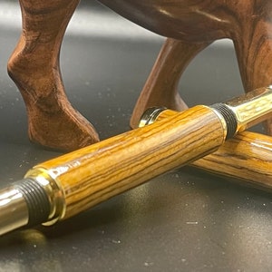 May include: A wooden pen with gold accents and a black tip. The pen is made of wood with a striped pattern.