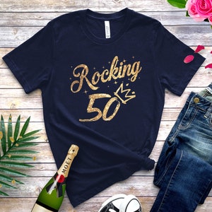 May include: A navy blue t-shirt with gold glitter text that says "Rocking 50" with a gold glitter crown above the 50. The shirt is laid out on a wooden surface with a bottle of champagne, a pair of jeans, and some rose petals.