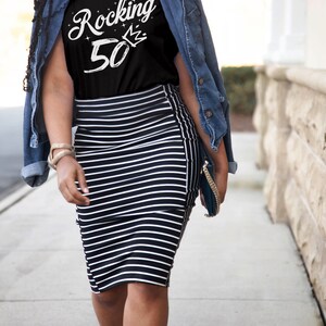 May include: A woman wearing a black t-shirt with the text "Rocking 50" in white with a crown above the 0. She is wearing a denim jacket and a black and white striped skirt.