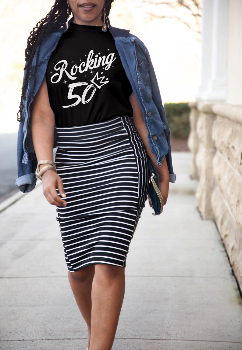 May include: A woman wearing a black t-shirt with the text "Rocking 50" in white with a crown above the 0. She is wearing a denim jacket and a black and white striped skirt.