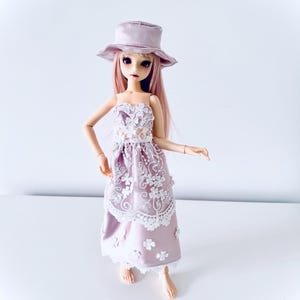 Può includere: Bambola con un vestito lavanda con dettagli in pizzo floreale bianco e un cappello abbinato. La bambola ha lunghi capelli rosa e si trova su una superficie bianca. L'abito ha un corpetto senza spalline e una gonna fluida.