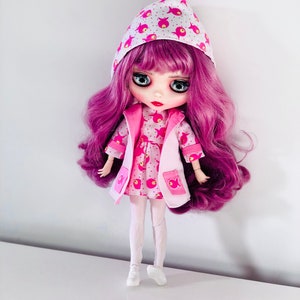 Può includere: Una bambola rosa e bianca con lunghi capelli viola che indossa un vestito rosa e bianco con un motivo a pesce e una giacca bianca con cappuccio con pesci rosa.