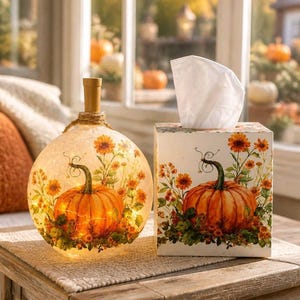 May include: A spherical glass light with a cork stopper and a square tissue box cover, both decorated with a matching print of an orange pumpkin surrounded by orange and yellow flowers. The glass light glows with internal warm white fairy lights.