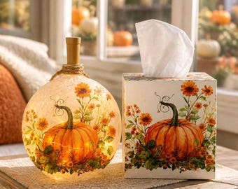 Puede incluir: Una lámpara esférica de cristal con tapón de corcho y una caja de pañuelos cuadrada, ambas decoradas con un estampado a juego de una calabaza naranja rodeada de flores naranjas y amarillas. La lámpara de cristal emite una luz cálida.