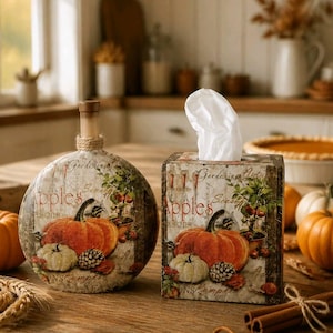 May include: A decorative bottle and tissue box set with a rustic autumn theme. The bottle has a wooden stopper and is adorned with a pumpkin design. The tissue box features a similar pumpkin and leaf motif, with the word "Apples" visible.