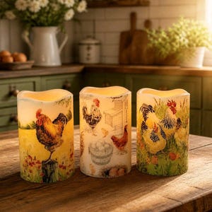 May include: Three cylindrical LED candles featuring farm-themed illustrations. The left candle shows a rooster on a post, the center displays a coop with eggs in a basket, and the right candle depicts a group of chickens in a grassy field.