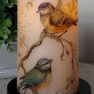 May include: A cylindrical pillar candle with a cream-colored wax surface featuring a printed illustration of two birds perched on branches, accented by faint script text. The candle is mounted on a solid black circular base and labeled with #1.
