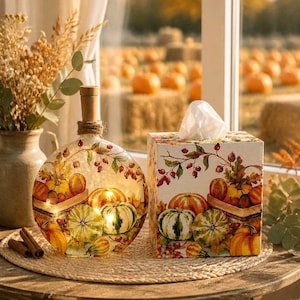 May include: A round glass bottle with a cork stopper and a square tissue box cover, both decorated with a colorful autumn print of pumpkins, berries, and leaves. The glass bottle features internal warm lighting and a jute twine neck wrap.
