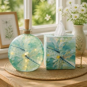May include: A round glass bottle lamp with a cork stopper and twine wrap, featuring a watercolor dragonfly and lily print with internal fairy lights, paired with a matching square tissue box cover decorated with the same dragonfly and lily motif.